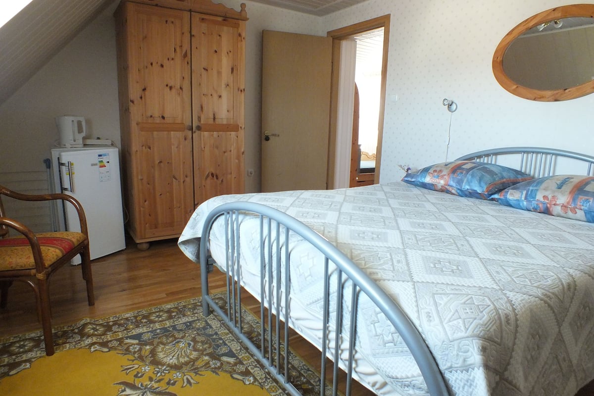 Ein Schlafzimmer mit einem Bett, einem Holzschrank, einem Sessel, einem kleinen Kühlschrank und einem Spiegel an der Wand.