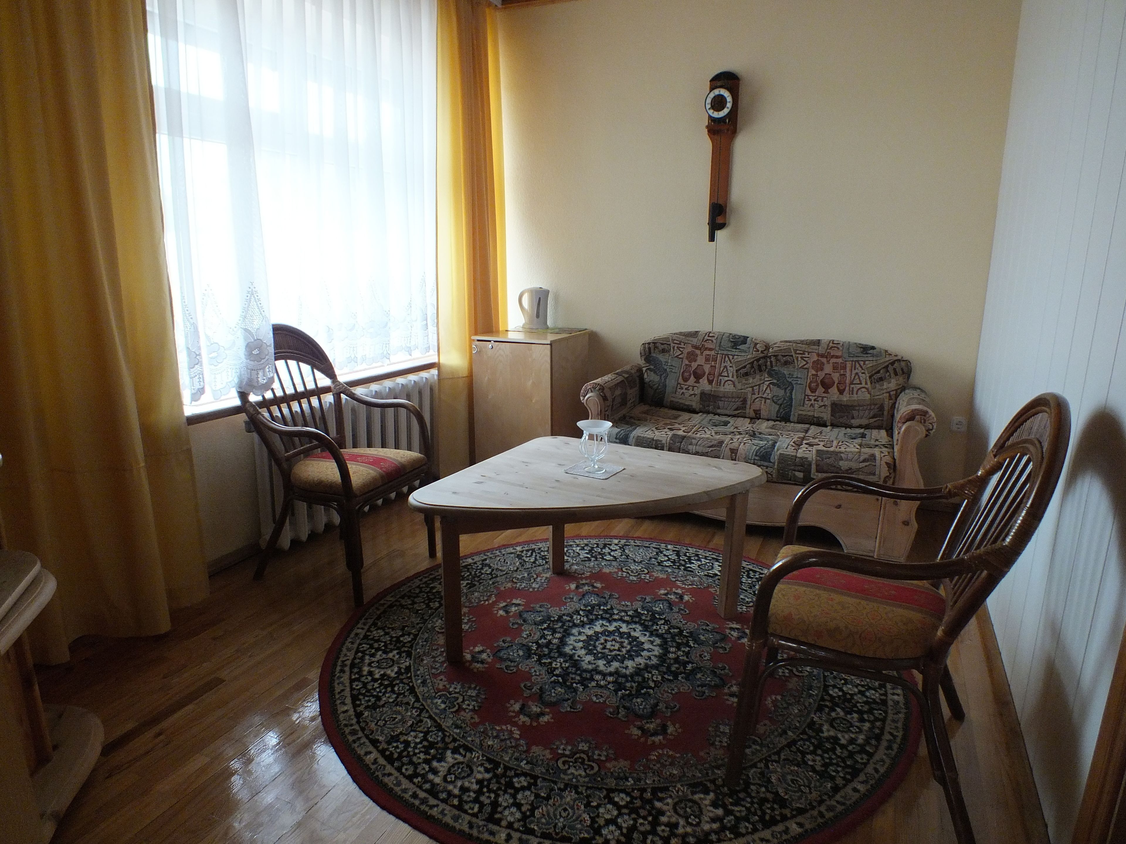 A cozy living room with a round wooden table, two wooden chairs, a patterned sofa, a radiator, and a wall clock.