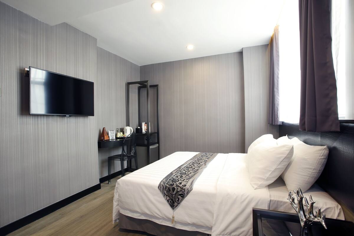 Een moderne hotelkamer met een flatscreen televisie aan de muur, een bed met witte lakens, een decoratief plaid en een bureau met een stoel en een koffiezetapparaat.