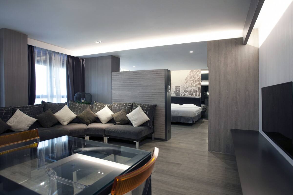 Een moderne woonkamer met een glazen salontafel, een bank met kussens, een bed en een flatscreen televisie.