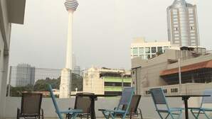 A balcony with chairs and a table, overlooking a cityscape with a prominent tower and a modern building. - 1000 Miles (Kuala Lumpur)
