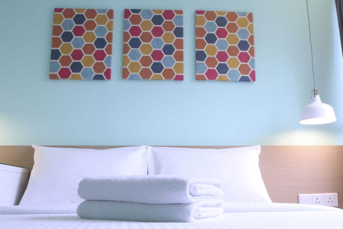 A hotel room with a bed, white pillows, a neatly folded white towel, and three hexagonal-patterned wall art pieces.