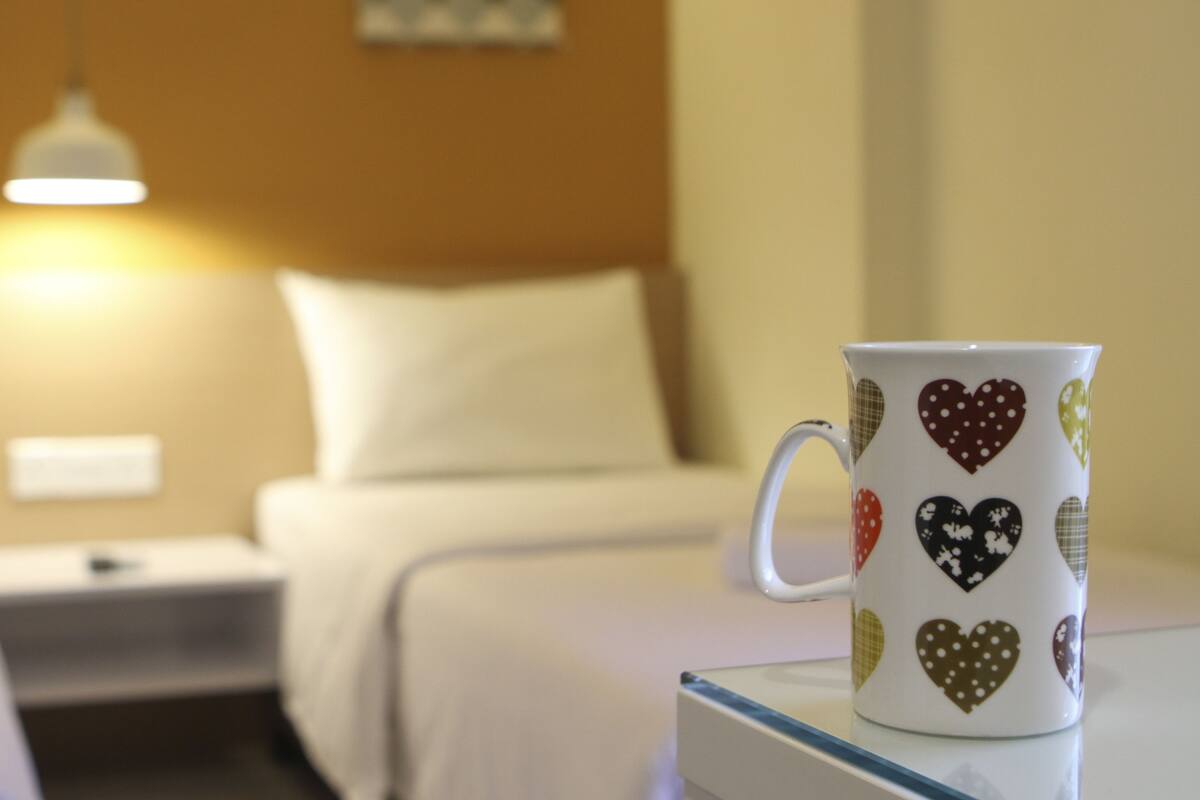 A hotel room with two beds, a nightstand, a lamp, and a decorative mug on a table.