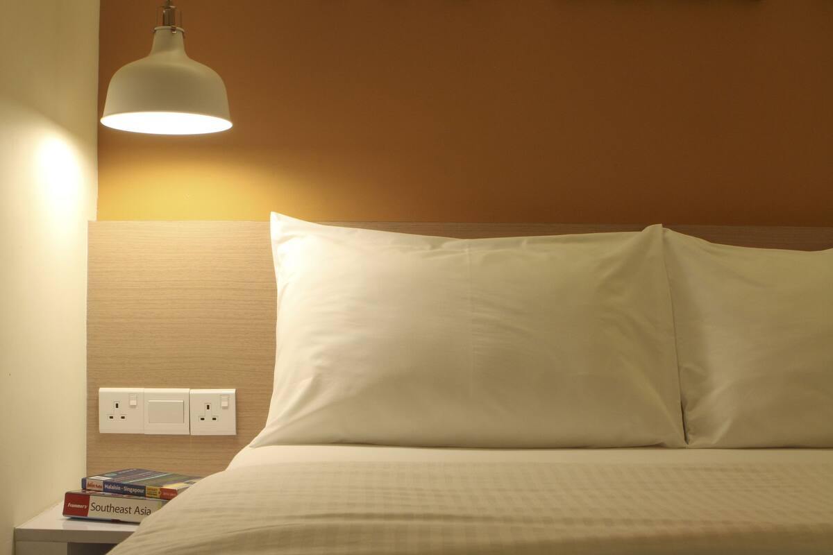A neatly made bed with white pillows and a white duvet, a bedside table with a lamp, and a wall socket.