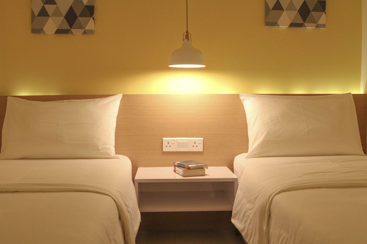 A hotel room with two single beds, a bedside table with a lamp, and a power outlet.