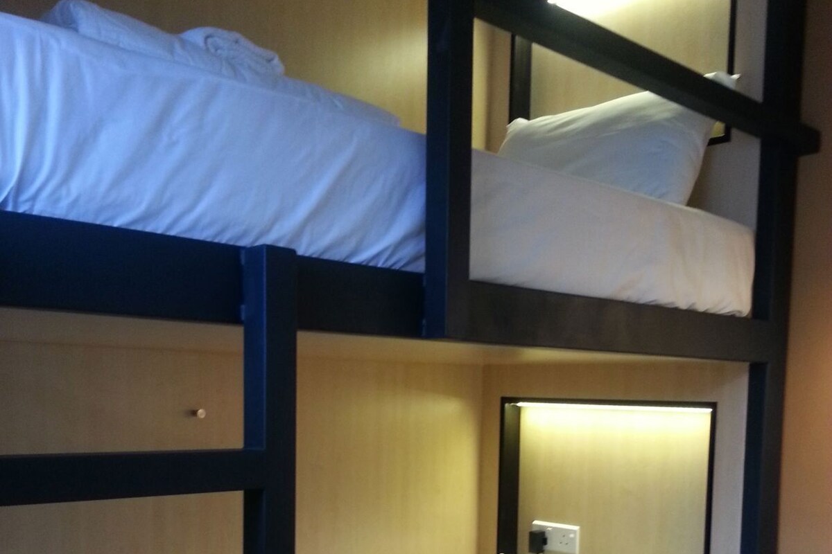 A bunk bed room with a desk and chair, a wall socket, and a window.