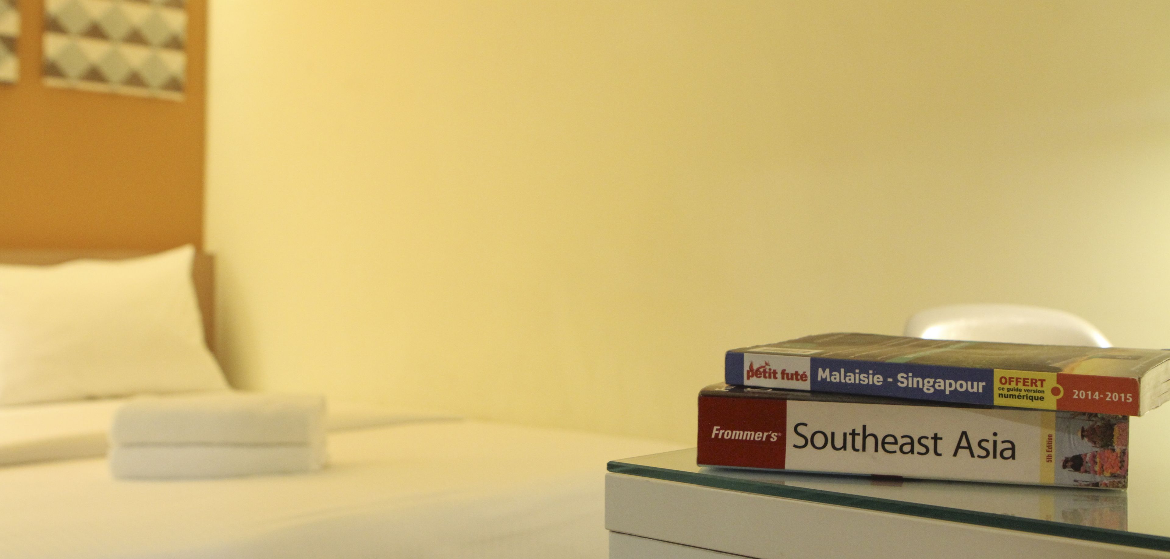 A bed with white linens and pillows, a glass table with a stack of travel guide books, and a white nightstand.