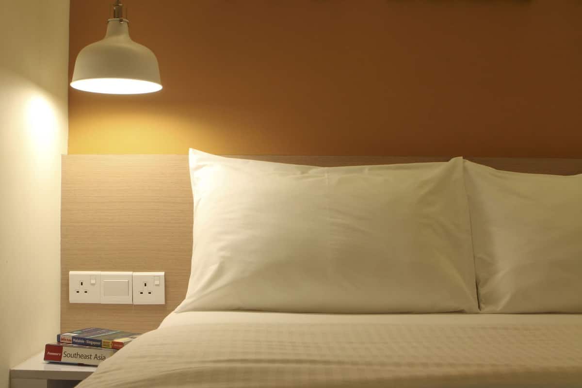 A neatly made bed with white bedding, a bedside lamp, and a nightstand with books and a power outlet.