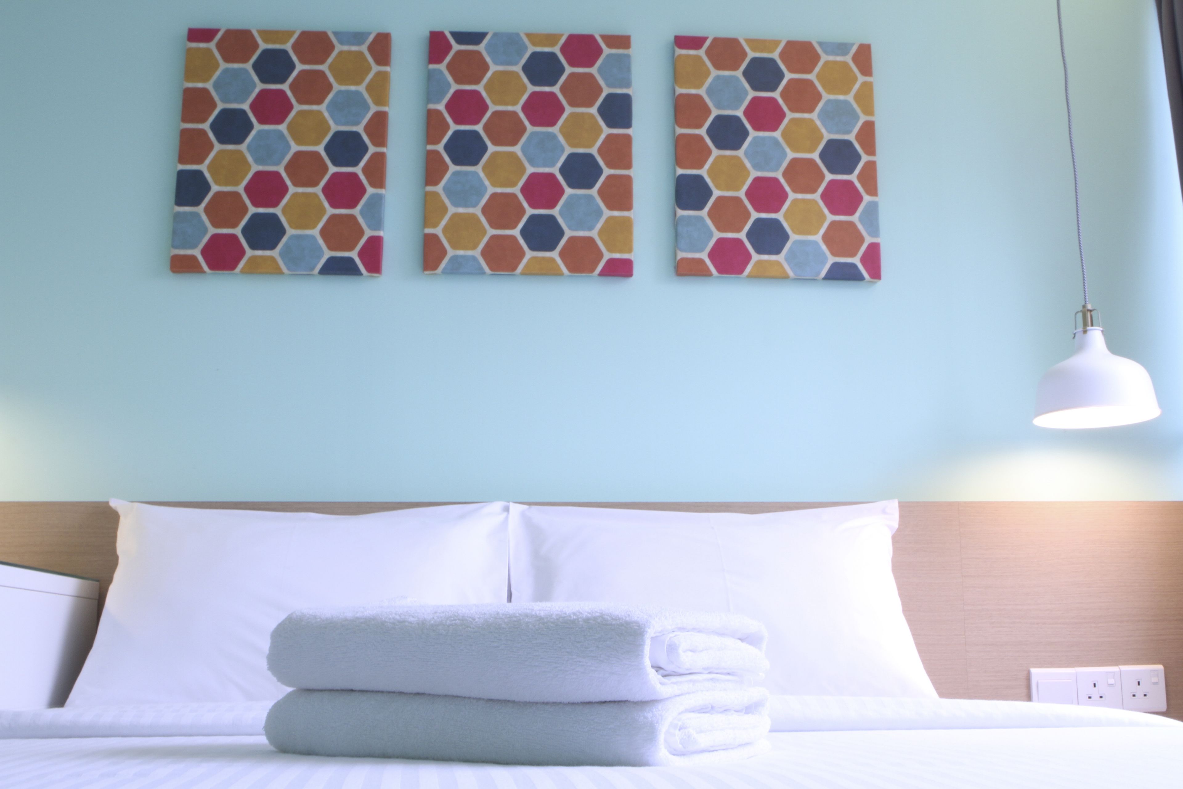 Un lit bien fait, avec du linge de lit blanc et deux serviettes pliées, un mur bleu clair et trois œuvres d’art aux motifs hexagonaux au-dessus du lit.
