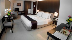 A hotel room with a bed, bedside table, desk, chair, and a small table with a vase of flowers. - Citystate Tower Hotel (Manila)