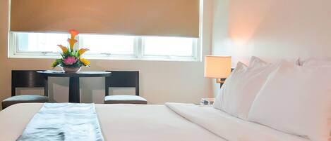 A hotel room with a large bed, a desk with a lamp, and a window with blinds.