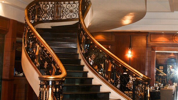 A grand spiral staircase with ornate black and gold railings, a large chandelier, and a polished wooden ceiling. - Citystate Tower Hotel (Manila)