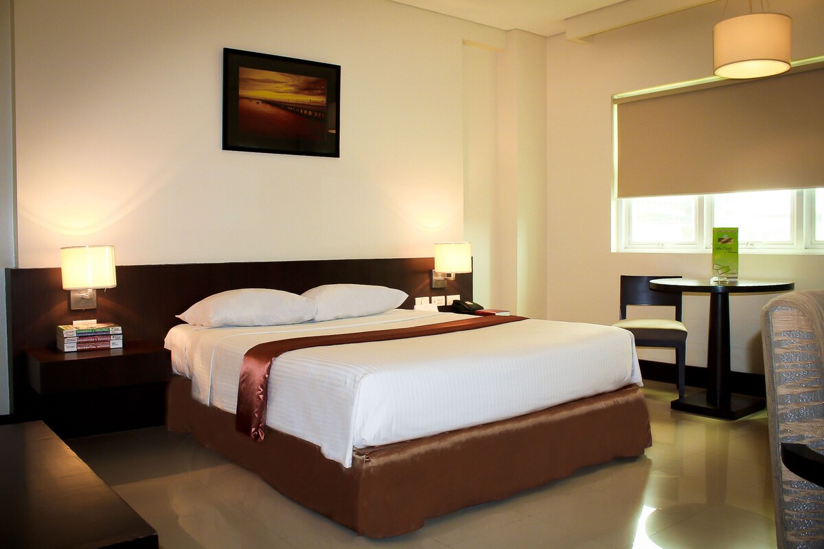 A hotel room with a large bed, two bedside lamps, a desk, a chair, and a window.