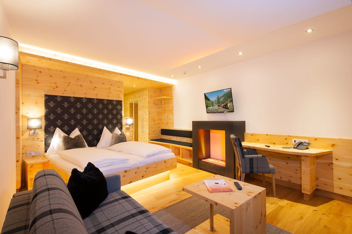 A modern hotel room with a bed, a desk, a chair, a small table, and a fireplace.