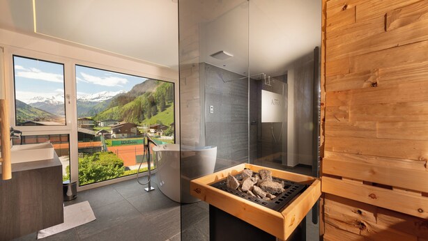 A modern bathroom with a glass shower enclosure, a wooden sauna, and a view of a mountainous landscape. - Hotel Alpina (Rauris)