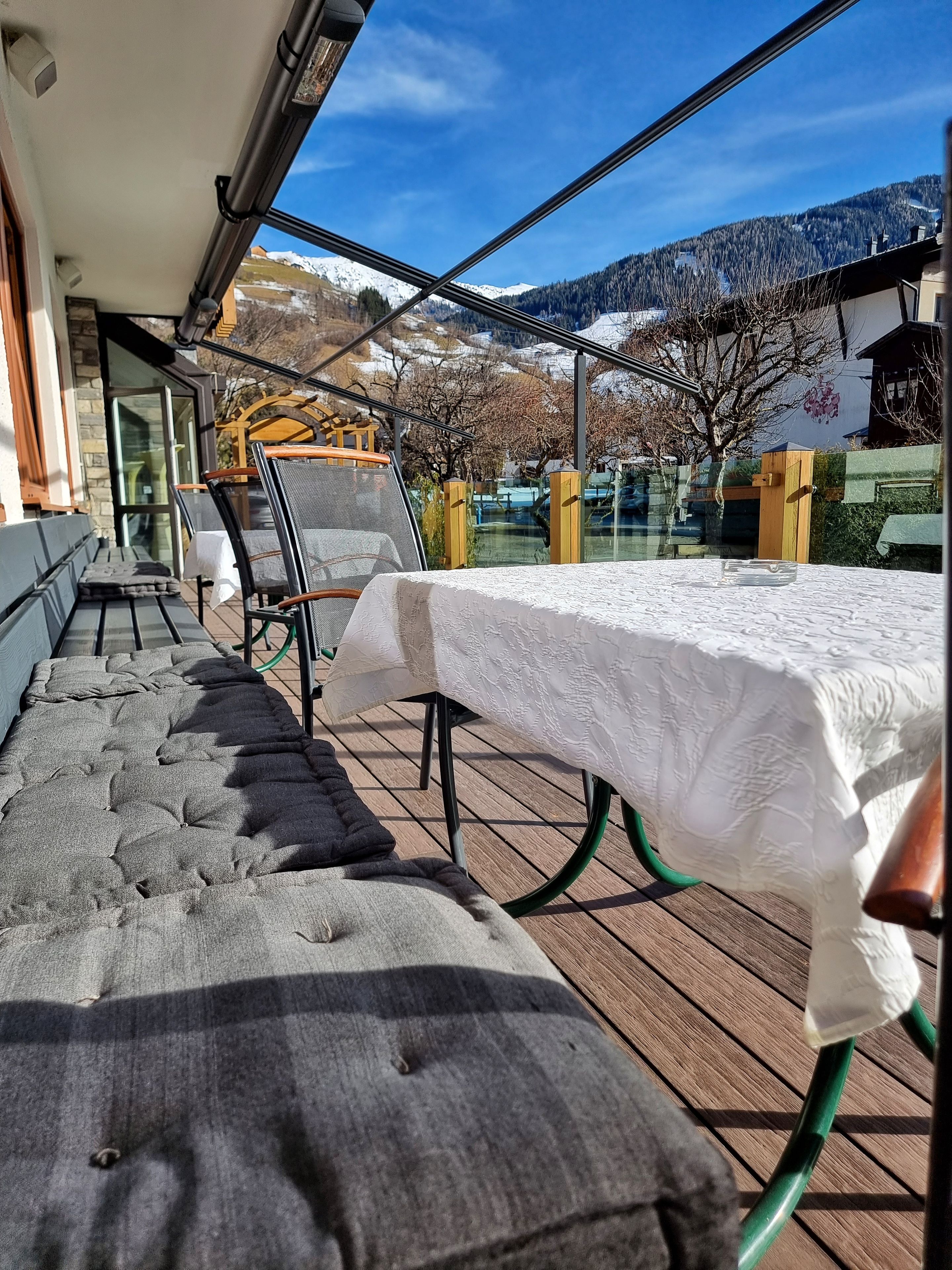 An outdoor seating area with a table covered in a white tablecloth, cushioned benches, and a view of snow-capped mountains.