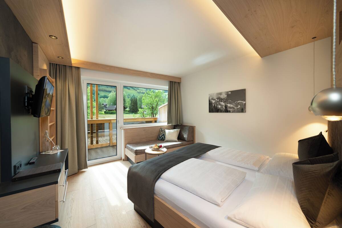 A modern hotel room with a large bed, a TV, a desk, and a balcony with a view of greenery.
