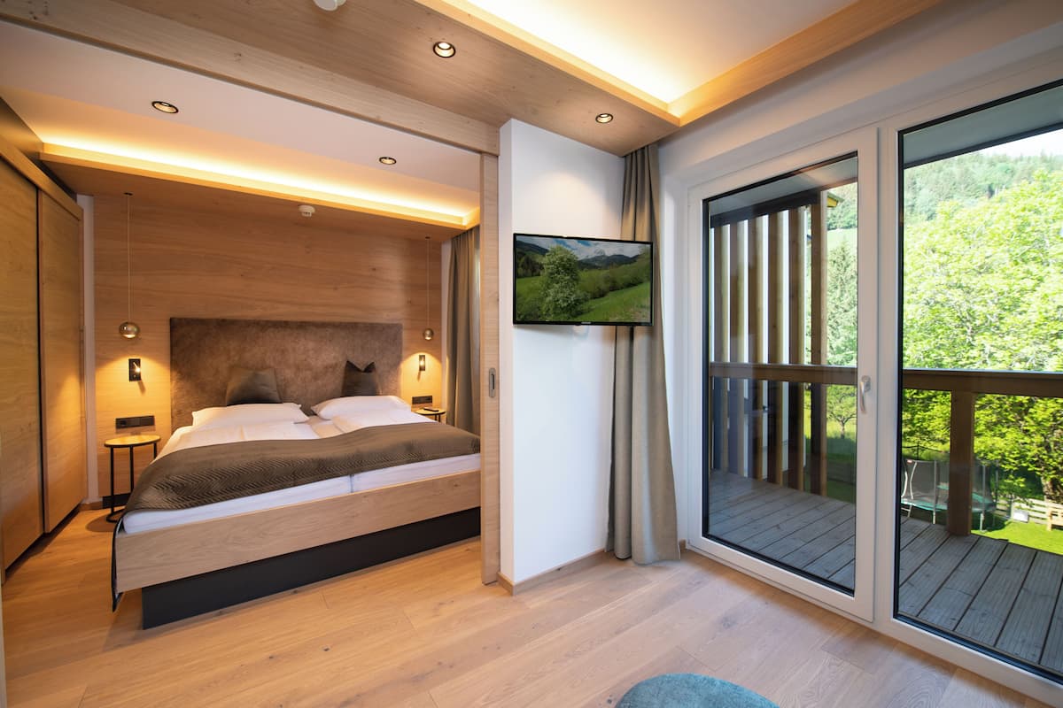 A modern bedroom with a large bed, a TV mounted on the wall, and a balcony with a view of greenery.