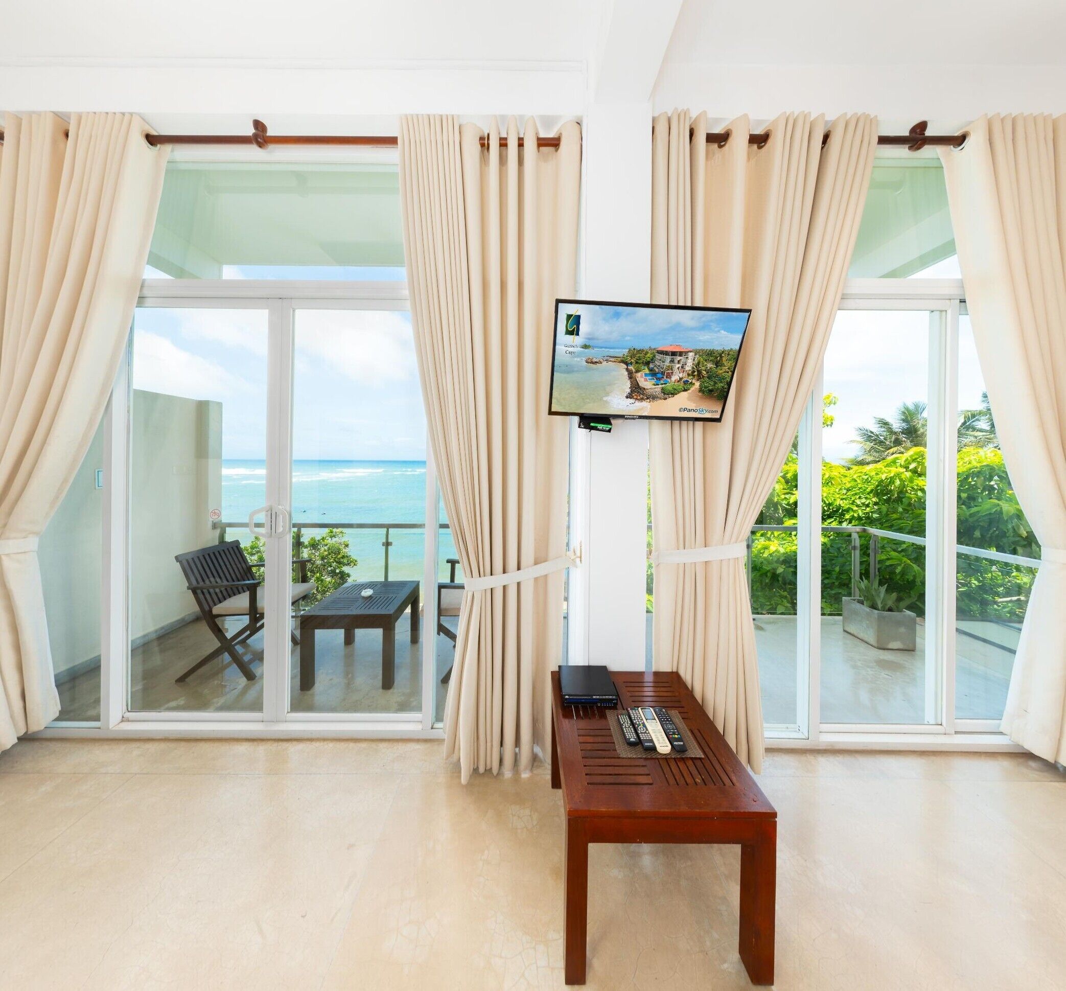 A room with a TV on the wall, a wooden table, and a view of the ocean through large glass doors.
