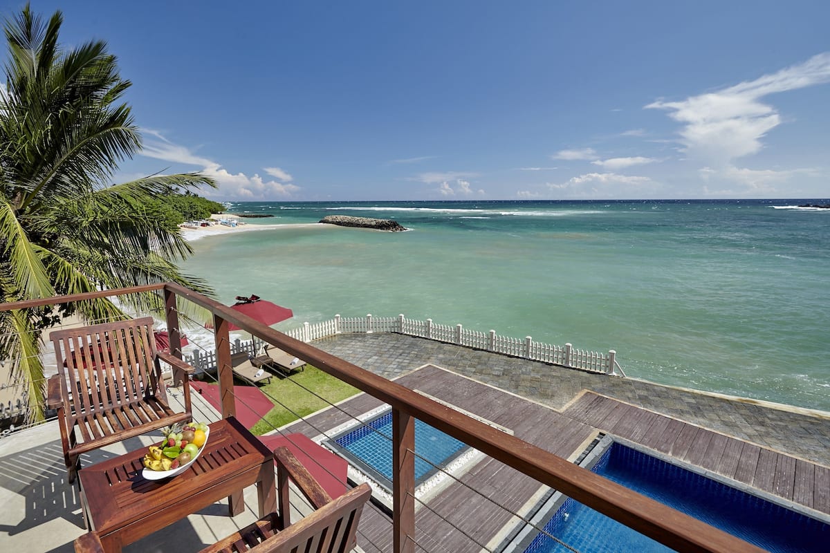 Una terraza de madera con piscina, con vista a una playa de aguas turquesas cristalinas y un promontorio rocoso.