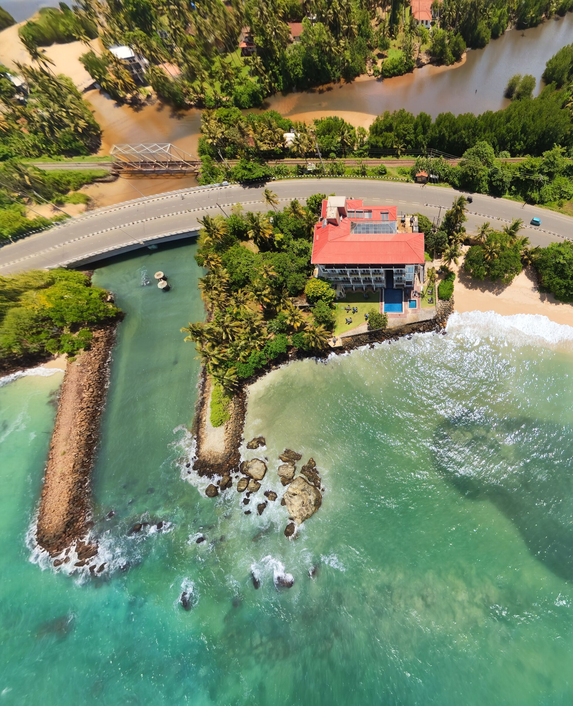 An aerial view of a coastal property with a beach, a small pier, and a building with a red roof.