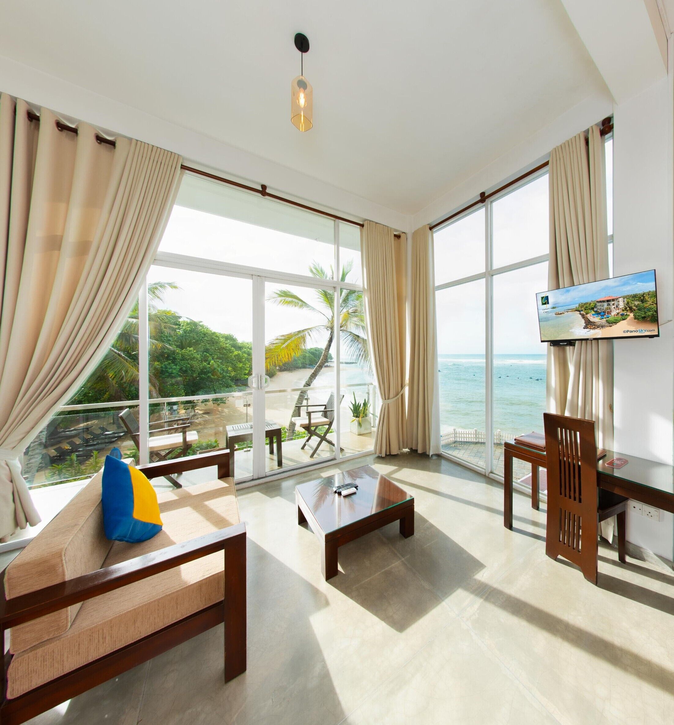 A spacious room with a large window offering a view of the beach and ocean, a comfortable seating area with a sofa and a coffee table, and a flat-screen TV mounted on the wall.