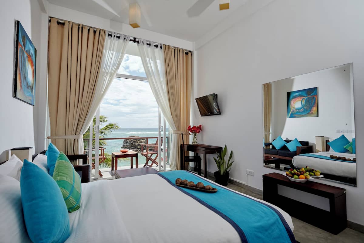 Habitación de hotel con cama, escritorio, silla, una pequeña mesa con fruta, planta en maceta, televisor y balcón con vista al océano.