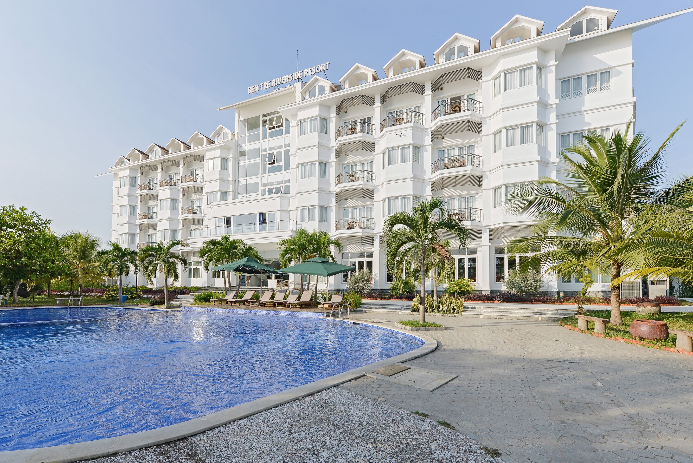 Ein mehrstöckiges Hotel mit Schwimmbad, Palmen und Liegestühlen.