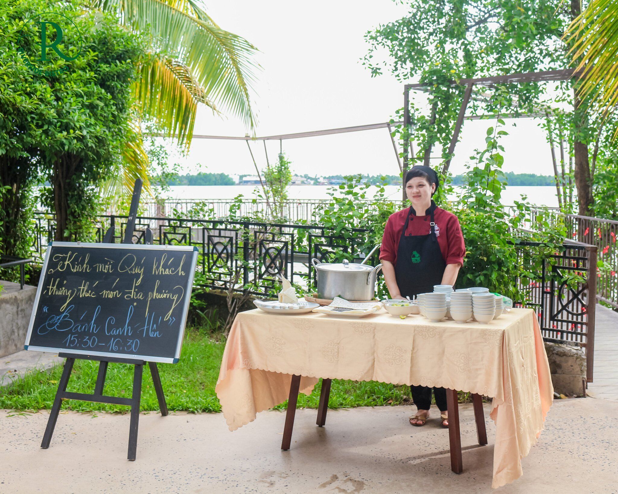 A person standing behind a table with dishes and a chalkboard sign, outdoors with greenery and a body of water in the background.