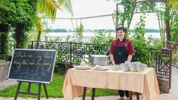 A person standing behind a table with dishes and a chalkboard sign, outdoors with greenery and a body of water in the background.