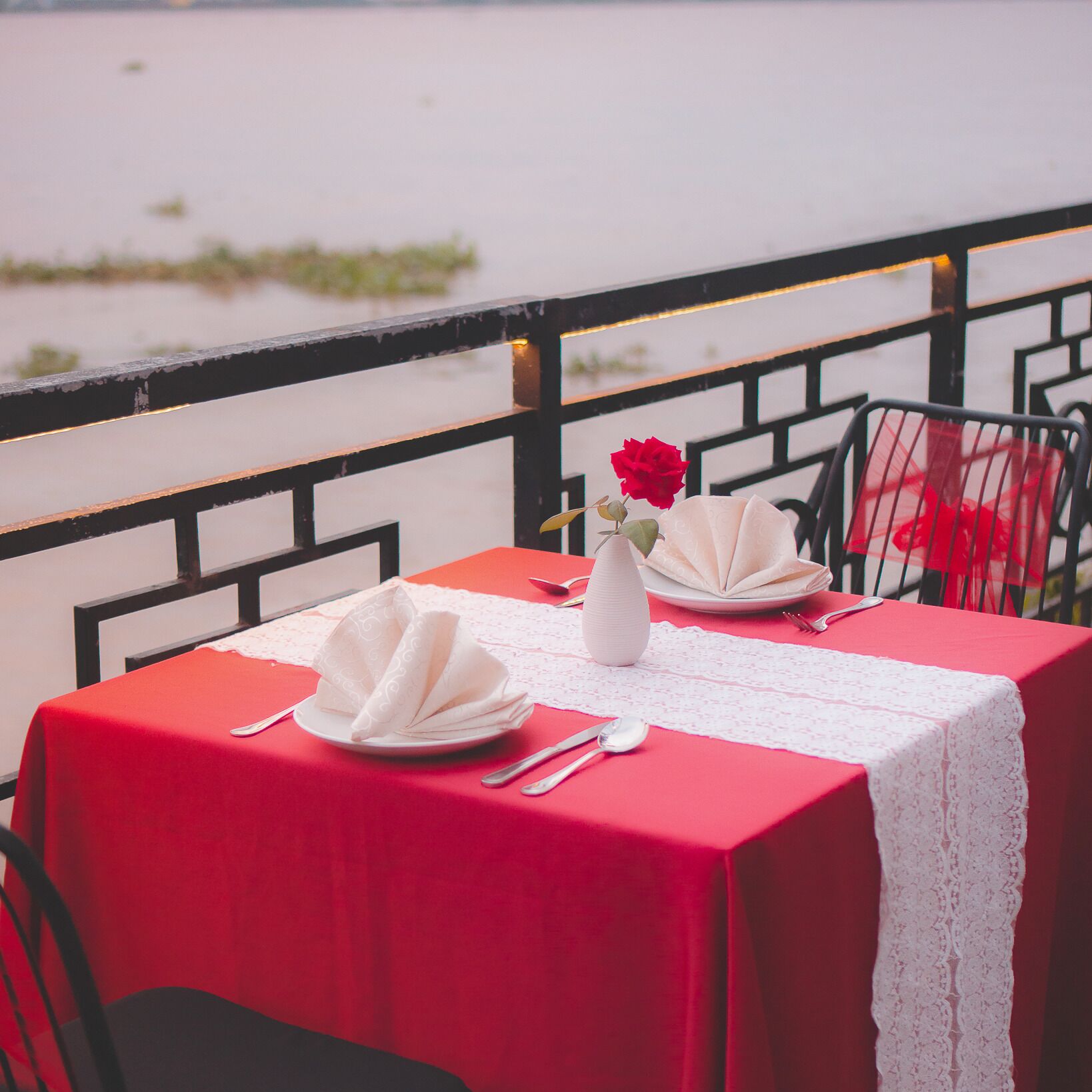 A table set for two with a red tablecloth, white napkins, a white vase with a red flower, and a view of a body of water.