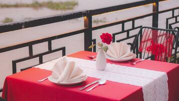 A table set for two with a red tablecloth, white napkins, a white vase with a red flower, and a view of a body of water.