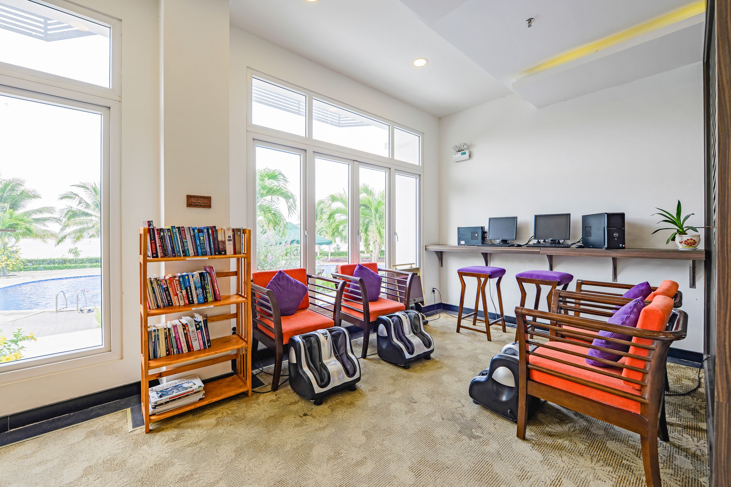 Un área de descanso moderna con estantería, sillones de masaje, escritorio con computadoras y grandes ventanales con vista a palmeras.