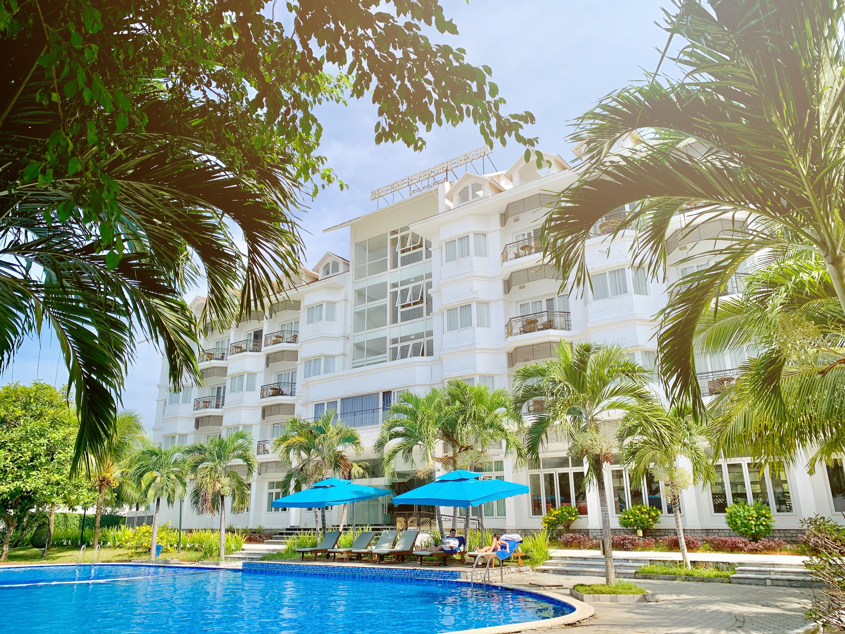 A hotel with a swimming pool surrounded by palm trees and blue umbrellas.