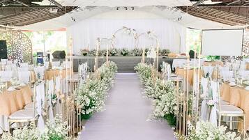 An indoor venue with a central aisle flanked by tables and chairs, a stage at the front, and a ceiling with a geometric pattern.