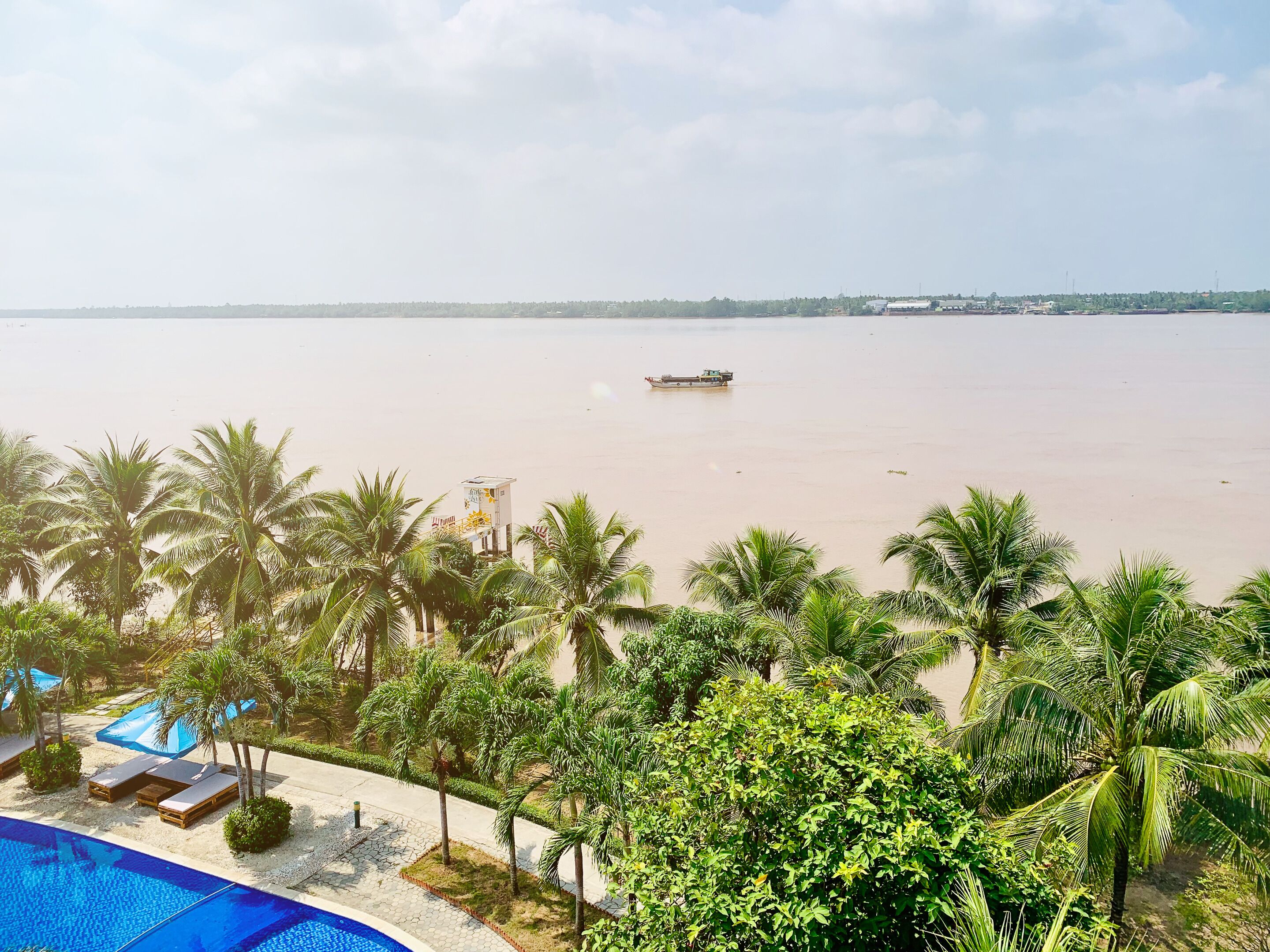 A poolside view with a boat on a calm body of water, surrounded by palm trees and a clear sky.