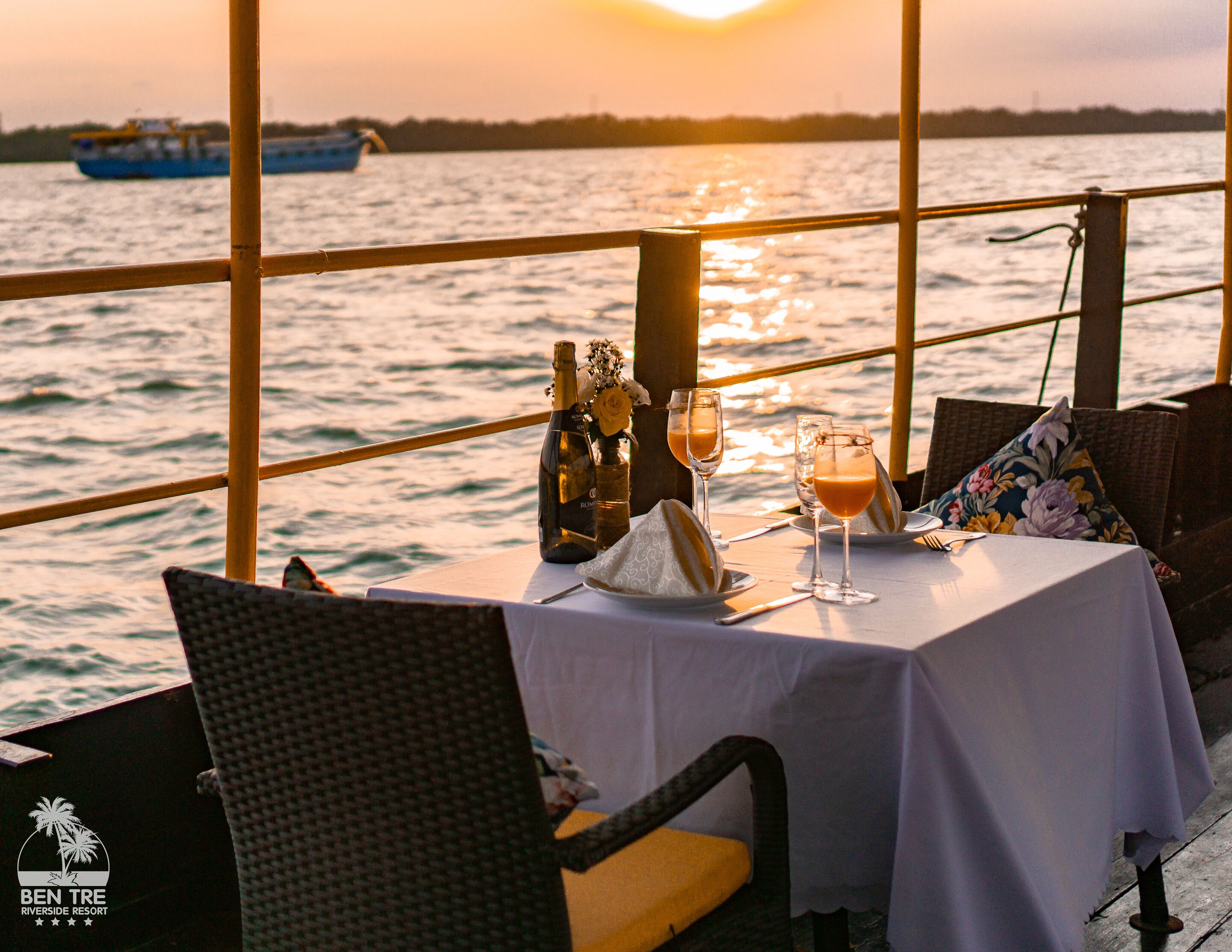 A table set for two with a bottle of champagne, two glasses, and a floral pillow on a table with a white tablecloth.