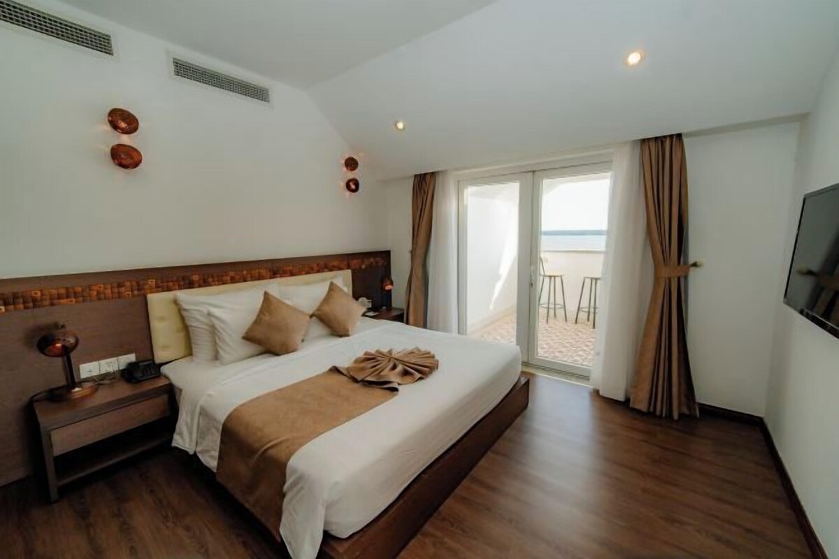 Quarto de hotel com uma cama grande, mesa de cabeceira, televisão e varanda com vista.