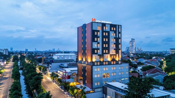 Un hotel moderno con un llamativo letrero de SwissExpress, rodeado de edificios urbanos y una calle arbolada.
