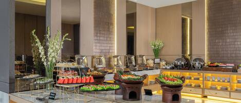 A modern hotel lobby with a buffet area featuring various food stations and a seating area with a decorative plant arrangement.