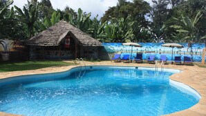 Une piscine entourée d’une terrasse en pierre, un petit chalet à toit de chaume et des chaises longues bleues.