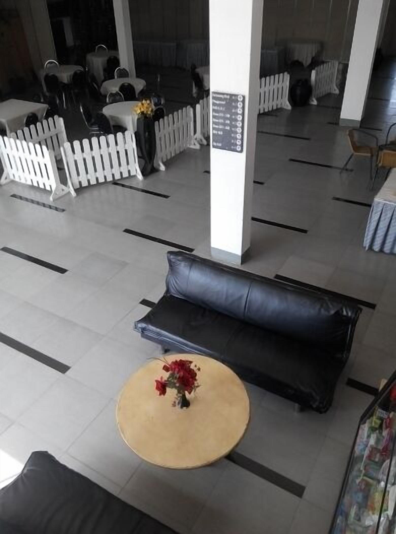 Un espace d’accueil avec un canapé en cuir noir, une table ronde de couleur beige ornée d’un vase de fleurs rouges et une clôture blanche en bois.