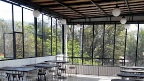 Un amplio espacio exterior con mesas y sillas, ventanas grandes y vista a árboles.