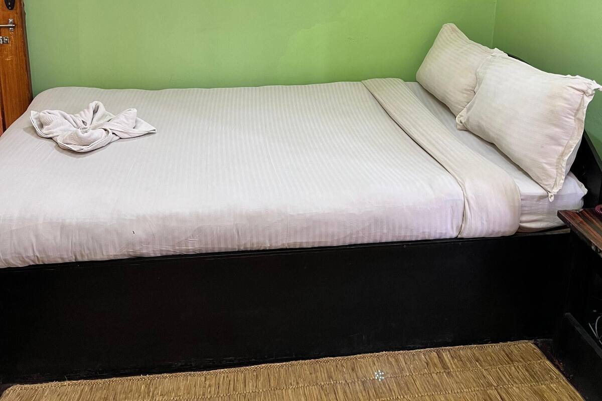 A single bed with a white mattress and pillows, a wooden headboard, a green wall, a framed painting, and a mirror with a curtain.