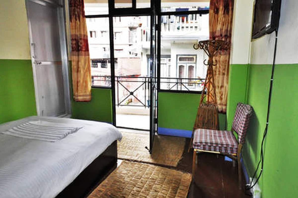 A room with a bed, a chair, a window with a view of buildings, and a balcony.