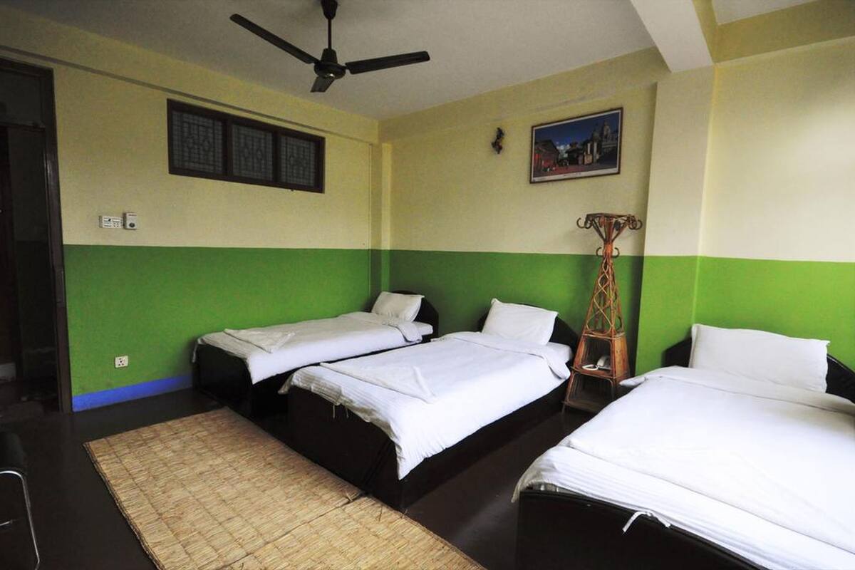 A room with three single beds, a ceiling fan, a picture on the wall, and a small table with a lamp.