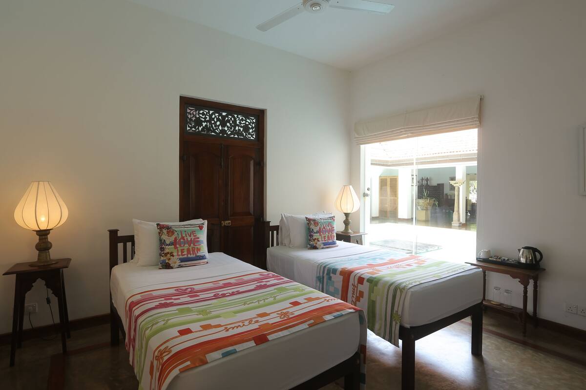 A room with two beds, each with a colorful striped bedspread and matching pillows. There are two bedside lamps, a wooden door with a decorative panel, and a window with a view of the outside.