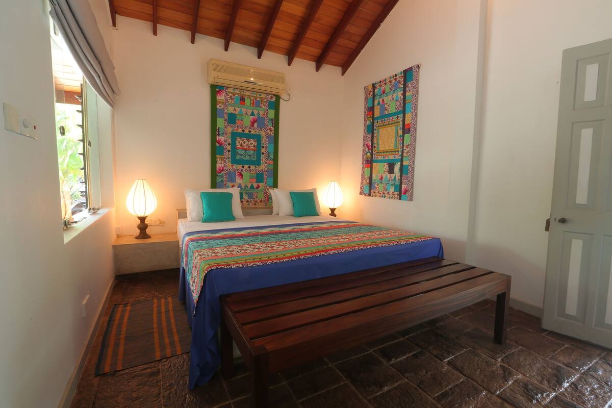 A bedroom with a wooden bed, two bedside lamps, a bench, a colorful quilt, and a quilted wall hanging.