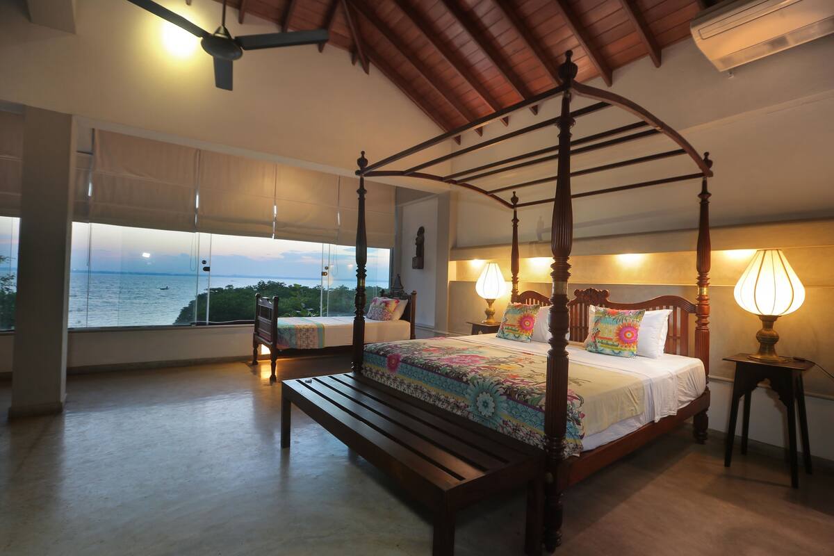 A four-poster bed with a view of the sea, a bench, and a bedside table with a lamp.