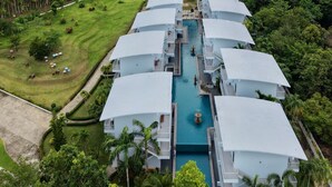 Un complexe hôtelier composé de plusieurs bâtiments aux toits blancs, disposés en rangée et reliés par une piscine centrale, le tout entouré d’une végétation luxuriante et dominé par un paysage montagneux.